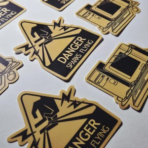Puede incluir: Una colección de pegatinas de estética retro. Las pegatinas presentan una combinación de colores negro y dorado. Una pegatina dice "DANGER SPARKS FLYING" con un gráfico de una persona y rayos. Otras pegatinas representan diseños de ordenadores antiguos.