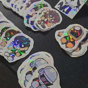 Puede incluir: Una colección de pegatinas holográficas con personajes de dibujos animados en varias poses. Las pegatinas tienen un acabado brillante e iridiscente y están dispuestas sobre una superficie oscura. Los diseños incluyen figuras con guitarras y otros personajes estilizados.