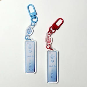 May include: Two acrylic keychains shaped like video game remote controllers. One features a light blue clasp and a charm with the number 1, while the other has a red clasp and a charm with the number 2. Both remotes display buttons and icons.
