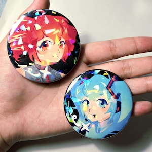 May include: Two circular pin-back buttons featuring anime-style character illustrations with a holographic shattered glass finish. One button displays a character with vibrant red hair, while the other features a character with light blue hair.