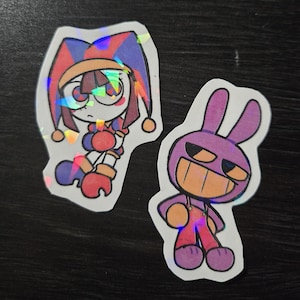 May include: Two holographic stickers featuring cartoon characters. One is a jester-like figure in red, blue, and orange, and the other is a purple rabbit-like character with a wide grin. Both stickers have a white border.