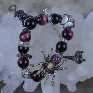 May include: A black and purple beaded bracelet with a silver spider charm and a butterfly charm. The bracelet is on a white background.