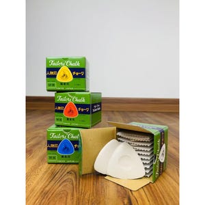 May include: Three boxes of tailor's chalk, each with a different colour: red, orange, and blue. The boxes are labelled "Tailors Chalk" in English and Japanese. The boxes are sitting on a wooden surface. There are also several white tailor's chalk triangles in a box.