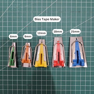 Può includere: Cinque strumenti per realizzare il nastro a sbieco di diverse dimensioni: 6 mm, 9 mm, 12 mm, 18 mm e 25 mm. Gli strumenti sono in metallo e hanno un'impugnatura in plastica. Le impugnature sono verdi, arancioni, gialle, rosse e blu.