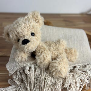 May include: A plush, beige teddy bear with black eyes and a black nose rests on a fringed, cream-colored blanket. The bear has soft, fluffy fur and is lying down. The blanket has a textured weave.