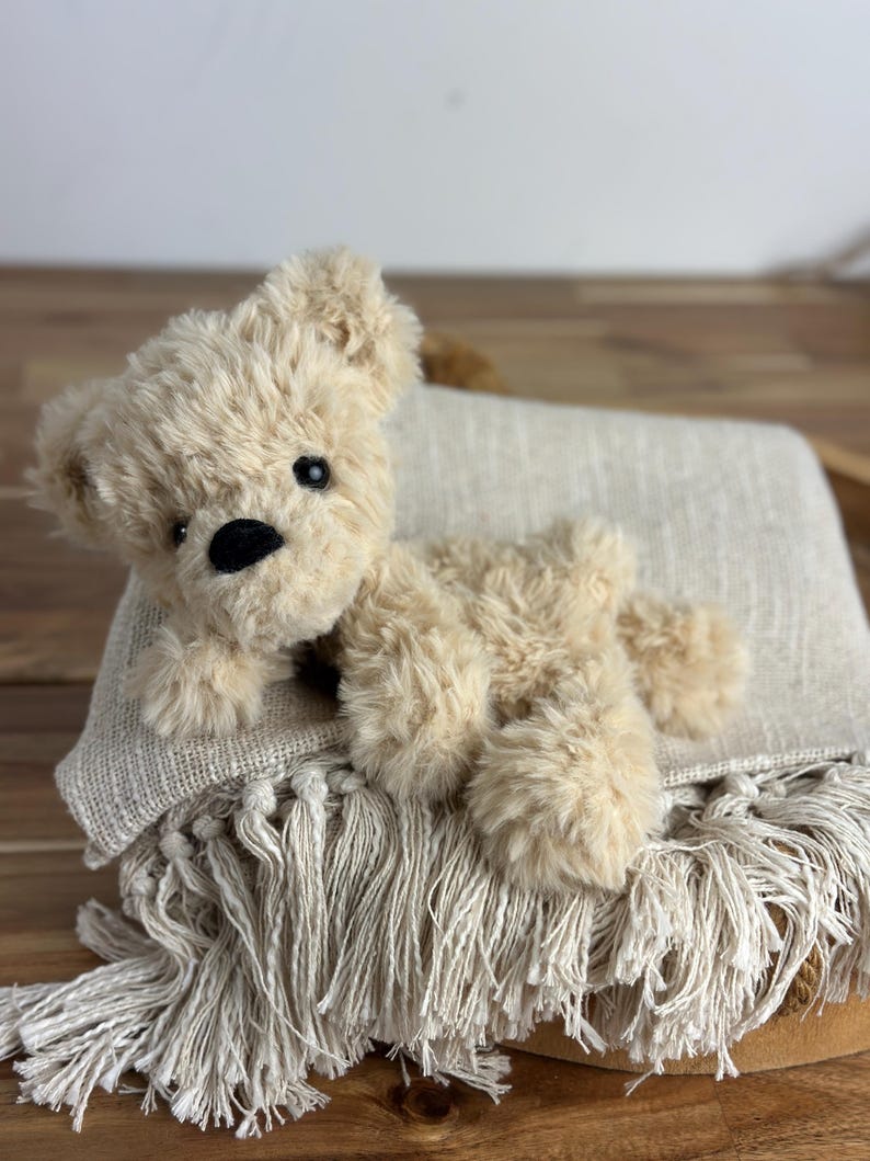 May include: A plush, beige teddy bear with black eyes and a black nose rests on a fringed, cream-colored blanket. The bear has soft, fluffy fur and is lying down. The blanket has a textured weave.
