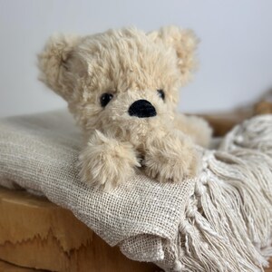 May include: A plush, beige teddy bear with a black nose and small black eyes rests on a textured, cream-colored blanket with fringe. The bear is lying down, with its paws visible. The background is a light, neutral color.