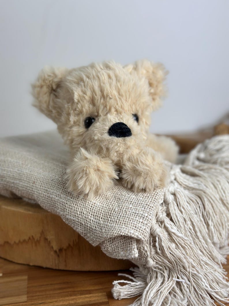 May include: A plush, beige teddy bear with a black nose and small black eyes rests on a textured, cream-colored blanket with fringe. The bear is lying down, with its paws visible. The background is a light, neutral color.