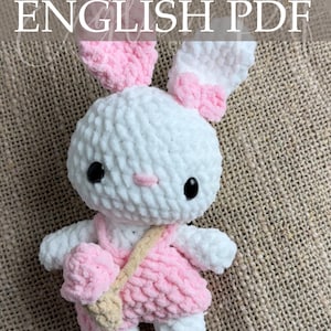 Op de afbeelding: Een handgemaakte witte en roze gehaakte konijnenpop met zwarte ogen, een roze neus en een roze jurk. De pop heeft een kleine beige tas en de tekst "ENGLISH PDF" is zichtbaar.