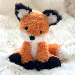 以下が含まれることがあります： オレンジ、白、黒の毛皮のぬいぐるみキツネ。キツネはふわふわのオレンジ色の体、白い胸と顔、黒い足と耳、白い先端の尾を持っています。おもちゃは白くふわふわした表面に座っています。