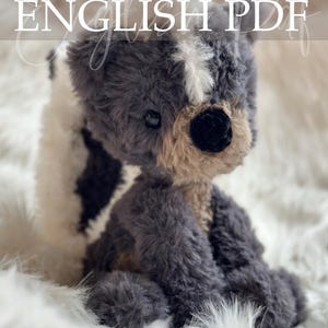 May include: A plush, handmade teddy bear in shades of gray, white, and tan. The bear has a black nose and is sitting on a fluffy white surface. The text "Crochet Pattern ENGLISH PDF" is overlaid on the image.