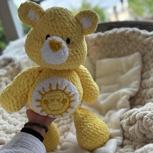 K&ouml;nnte beinhalten: Ein handgefertigter gelber H&auml;kelb&auml;r mit wei&szlig;en Akzenten an Ohren und Schnauze. Der B&auml;r hat ein l&auml;chelndes Sonnendesign auf dem Bauch. Das Kuscheltier ist weich und kuschelig, perfekt f&uuml;r ein Kinderzimmer oder als dekoratives Element.