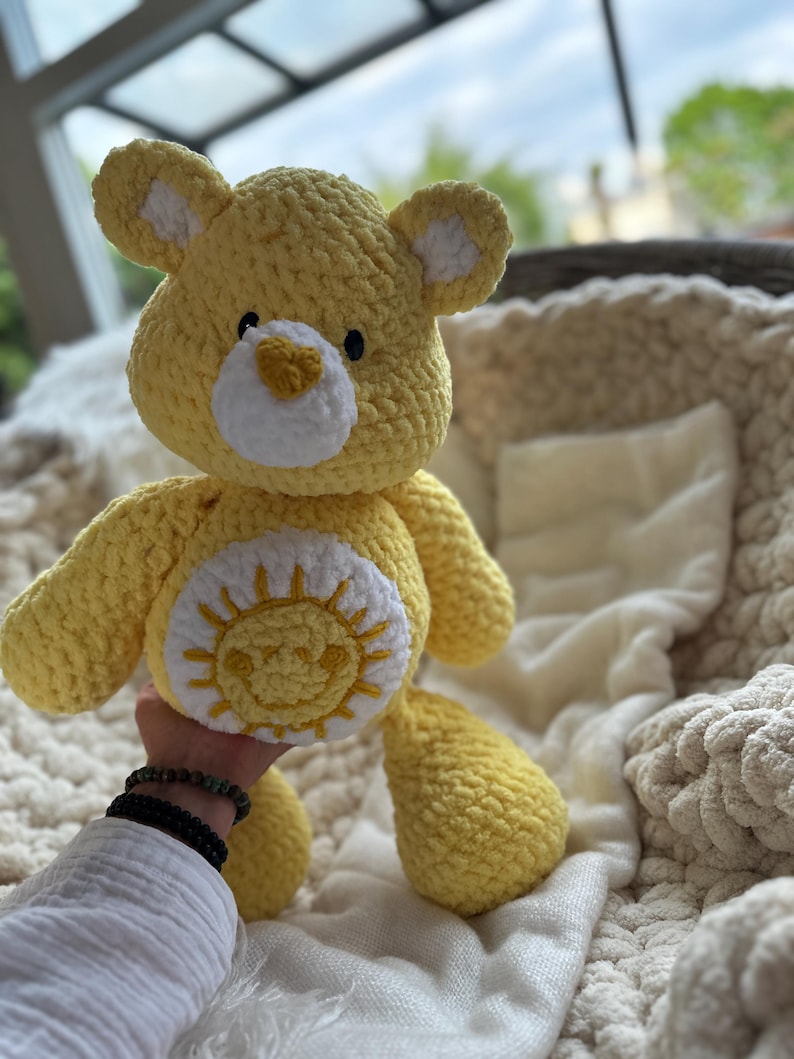 K&ouml;nnte beinhalten: Ein handgefertigter gelber H&auml;kelb&auml;r mit wei&szlig;en Akzenten an Ohren und Schnauze. Der B&auml;r hat ein l&auml;chelndes Sonnendesign auf dem Bauch. Das Kuscheltier ist weich und kuschelig, perfekt f&uuml;r ein Kinderzimmer oder als dekoratives Element.