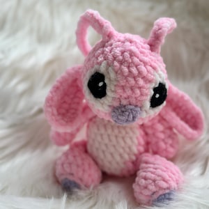 May include: A handmade crochet plush toy of a pink alien character with large ears, antennae, and a light pink belly. The figure features oversized black eyes with white highlights and a small purple nose, crafted from soft, textured yarn.