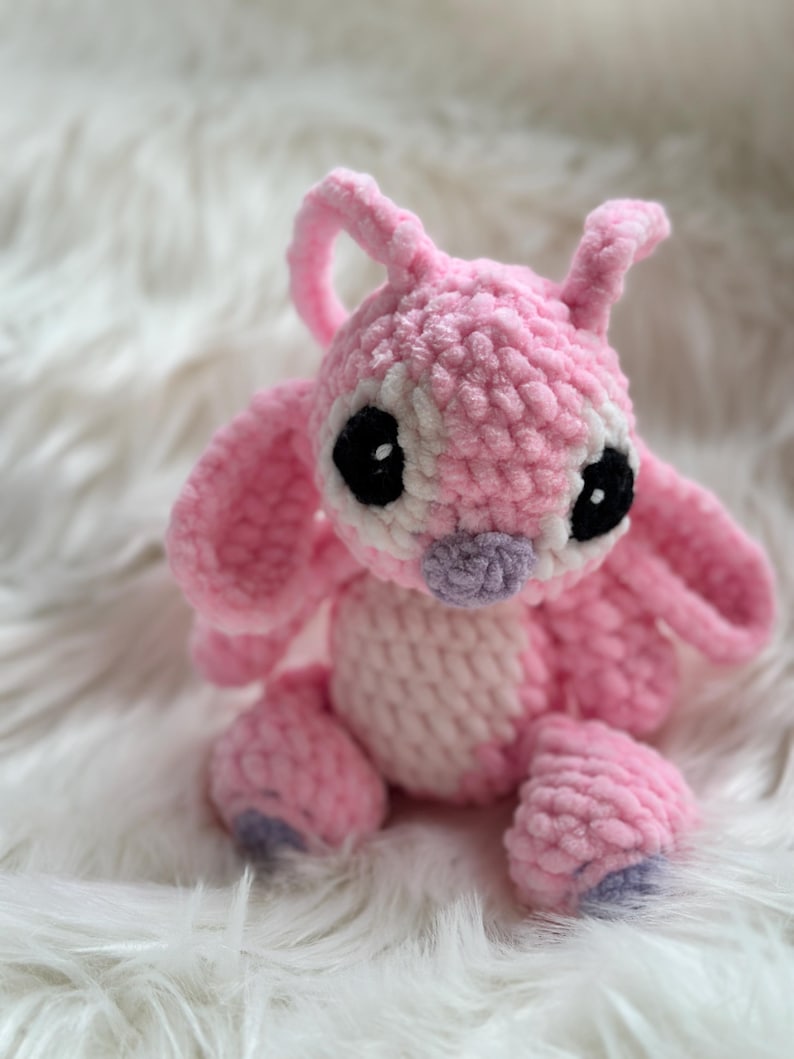 May include: A handmade crochet plush toy of a pink alien character with large ears, antennae, and a light pink belly. The figure features oversized black eyes with white highlights and a small purple nose, crafted from soft, textured yarn.