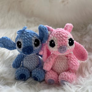 May include: Two handmade crochet plush toys featuring large ears and round black eyes. One figure is light blue with a darker blue nose and chest patch, while the other is light pink with a light purple nose and chest patch, both sitting upright.