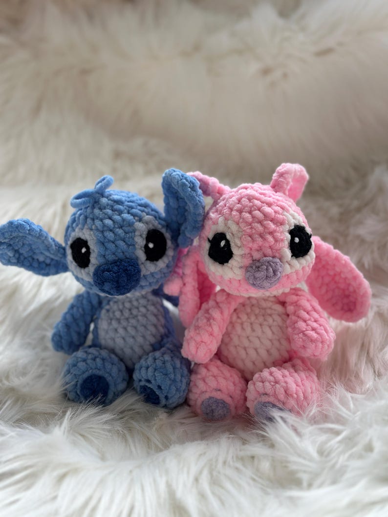 May include: Two handmade crochet plush toys featuring large ears and round black eyes. One figure is light blue with a darker blue nose and chest patch, while the other is light pink with a light purple nose and chest patch, both sitting upright.