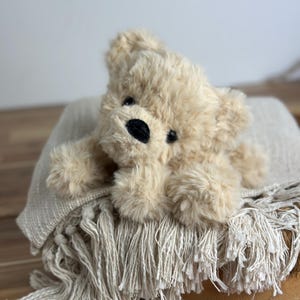 May include: A plush, beige teddy bear with a black nose and eyes rests on a fringed, cream-colored blanket. The bear is lying down, and the blanket is draped over a wooden surface, creating a cozy, inviting scene.