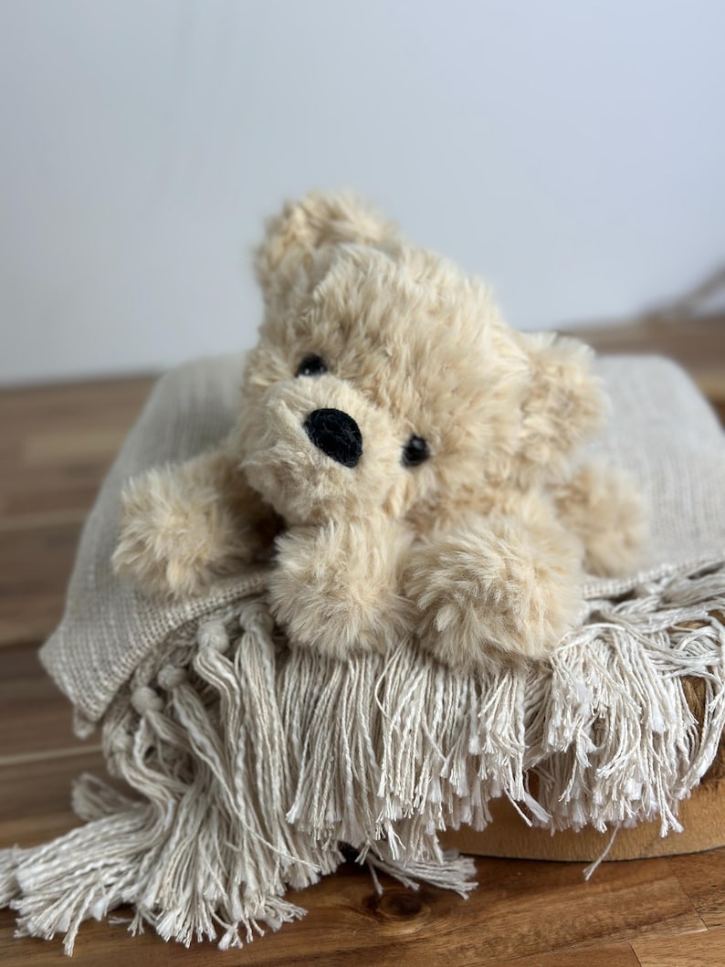 May include: A plush, beige teddy bear with a black nose and eyes rests on a fringed, cream-colored blanket. The bear is lying down, and the blanket is draped over a wooden surface, creating a cozy, inviting scene.