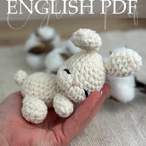 Op de afbeelding: Een crèmekleurig gehaakt lam met zwarte ogen en een roze neus wordt in een hand gehouden. Het lam heeft grote, pluizige oren en een kleine staart. De afbeelding bevat de tekst "Crochet Pattern ENGLISH PDF".