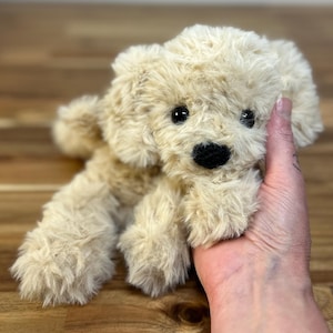 May include: A plush, beige puppy dog toy with a fluffy texture. The toy has black eyes and a black nose. The dog is lying down, and its paws are visible. The toy is held in a person's hand.