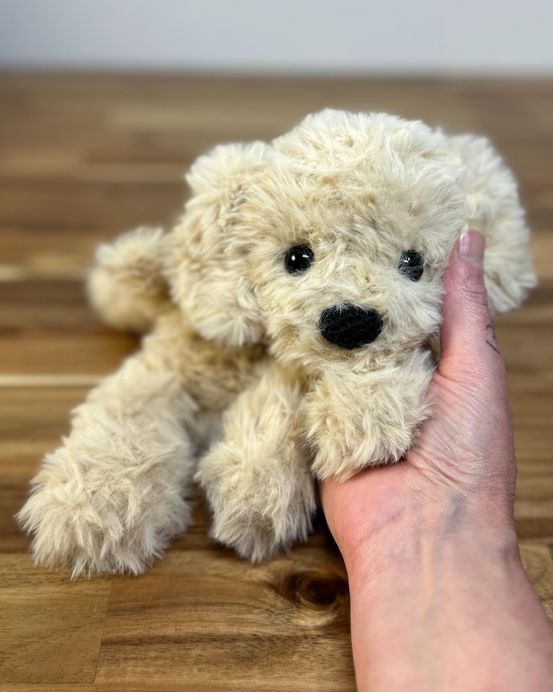 May include: A plush, beige puppy dog toy with a fluffy texture. The toy has black eyes and a black nose. The dog is lying down, and its paws are visible. The toy is held in a person's hand.