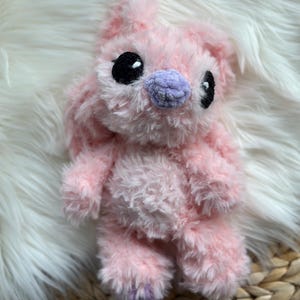 May include: A plush toy crafted from soft, light pink fuzzy yarn. The creature features large, round black eyes and a small, rounded lavender nose. It has long, floppy ears extending upwards and a rounded body with short arms and legs.