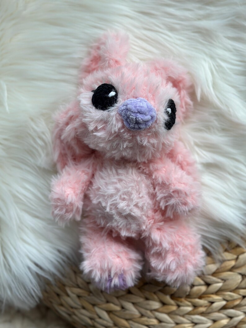 May include: A plush toy crafted from soft, light pink fuzzy yarn. The creature features large, round black eyes and a small, rounded lavender nose. It has long, floppy ears extending upwards and a rounded body with short arms and legs.