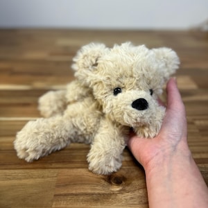 May include: A small, plush teddy bear in a light beige color. The bear has a black nose and black eyes. The fur is soft and fluffy. The bear is lying down on a wooden surface.