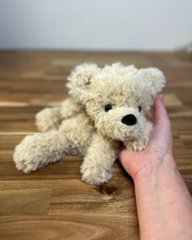 May include: A small, plush teddy bear in a light beige color. The bear has a black nose and black eyes. The fur is soft and fluffy. The bear is lying down on a wooden surface.