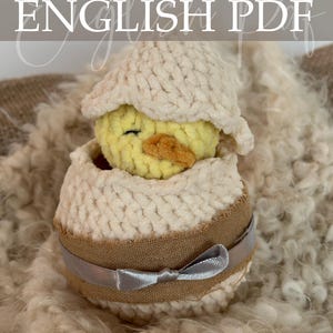 Op de afbeelding: Een gehaakt kuiken in een eierschaal, met de tekst "Crochet Pattern ENGLISH PDF". Het kuiken is geel met een oranje snavel en de eierschaal is crèmekleurig met een bruine band en een zilveren lint.