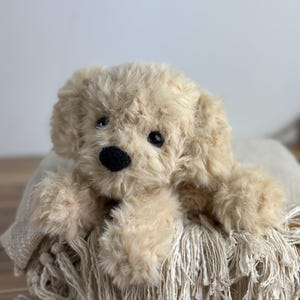 May include: A plush, beige-colored puppy dog toy with a black nose and dark eyes. The stuffed animal is lying on a fringed blanket. The toy has soft, fluffy fur and is designed to resemble a real dog.