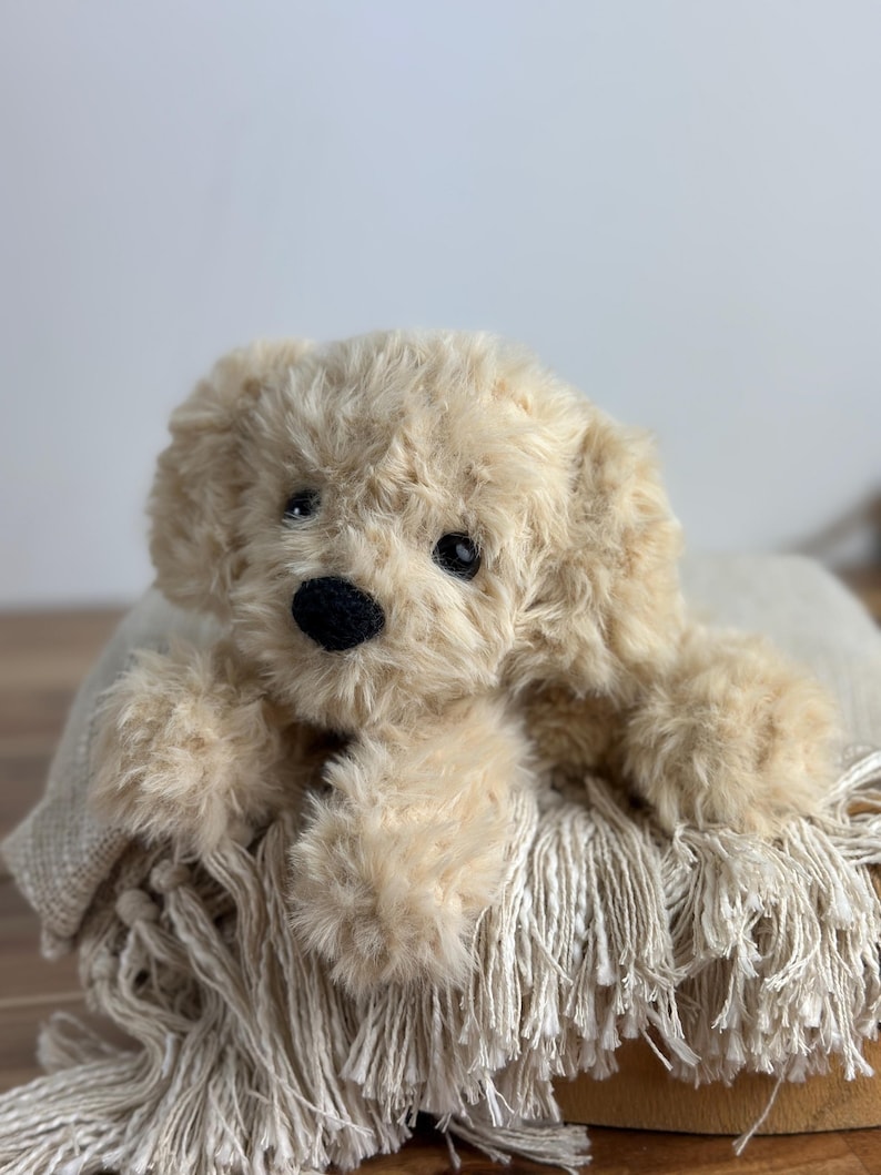 May include: A plush, beige-colored puppy dog toy with a black nose and dark eyes. The stuffed animal is lying on a fringed blanket. The toy has soft, fluffy fur and is designed to resemble a real dog.
