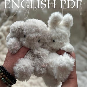 May include: Two plush, fluffy stuffed animals, an elephant and a bunny, are held in a person's hands. The toys are white and gray, with small black eyes. The text "Crochet Pattern ENGLISH PDF" is at the top of the image.