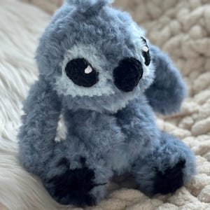 May include: A plush, fuzzy, blue-gray stuffed creature with a rounded head, large black oval eyes, a small black nose, and dark black patches on its feet. The toy is crafted from a soft, textured yarn material with a dense, shaggy appearance.