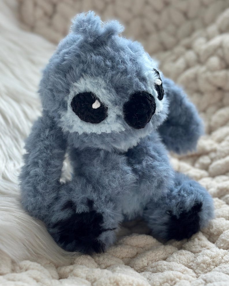 May include: A plush, fuzzy, blue-gray stuffed creature with a rounded head, large black oval eyes, a small black nose, and dark black patches on its feet. The toy is crafted from a soft, textured yarn material with a dense, shaggy appearance.