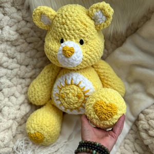 K&ouml;nnte beinhalten: Ein handgefertigter gelber H&auml;kelb&auml;r mit wei&szlig;en Akzenten an Ohren und Schnauze. Der B&auml;r hat einen wei&szlig;en Bauch mit einem gelben Sonnendesign und ein gelbes Herz auf der Pfote. Der B&auml;r sitzt auf einer wei&szlig;en, flauschigen Oberfl&auml;che.