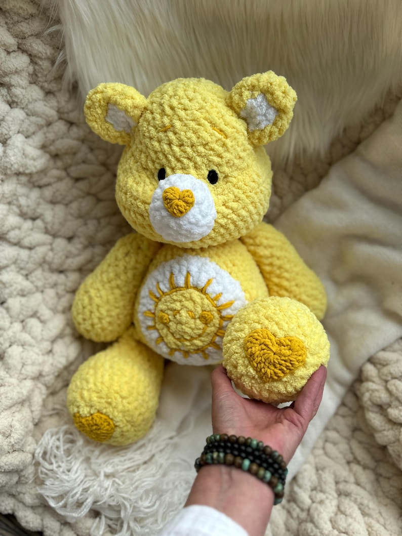 K&ouml;nnte beinhalten: Ein handgefertigter gelber H&auml;kelb&auml;r mit wei&szlig;en Akzenten an Ohren und Schnauze. Der B&auml;r hat einen wei&szlig;en Bauch mit einem gelben Sonnendesign und ein gelbes Herz auf der Pfote. Der B&auml;r sitzt auf einer wei&szlig;en, flauschigen Oberfl&auml;che.