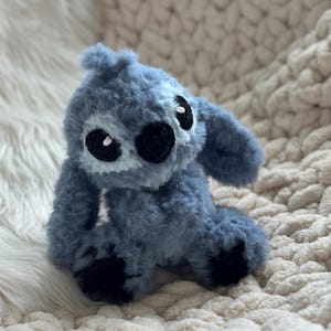 May include: A plush toy crafted from fuzzy blue yarn, featuring large black embroidered eyes, a prominent black nose, and long ears. The character has a textured, soft appearance with dark blue accents on the feet and a light blue patch on the face.