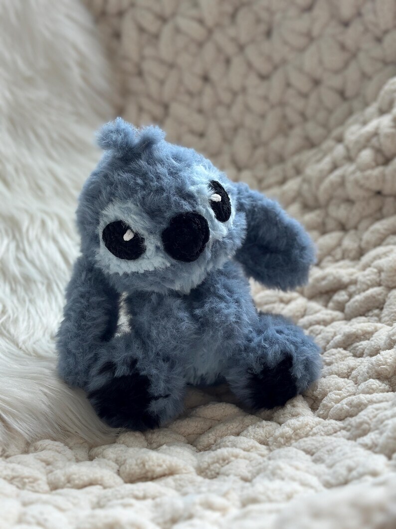 May include: A plush toy crafted from fuzzy blue yarn, featuring large black embroidered eyes, a prominent black nose, and long ears. The character has a textured, soft appearance with dark blue accents on the feet and a light blue patch on the face.