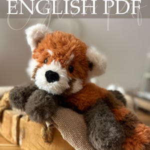 May include: A crocheted red panda soft toy with a fluffy texture. The toy features a combination of brown, orange, and white yarn, with a black nose and small black eyes. The image includes the text "Crochet Pattern ENGLISH PDF" at the top.