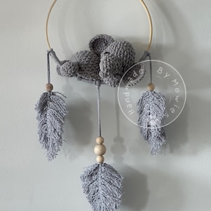 Puede incluir: Un elefante de crochet gris está colgado de un aro de madera con plumas grises y blancas y cuentas de madera. El aro tiene un círculo blanco con el texto "Handmade by Mollies"
