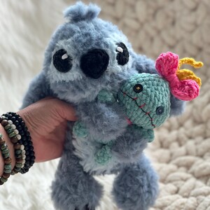 May include: A fuzzy blue crochet plush toy resembling a koala-like creature with large black eyes and a black nose, holding a smaller light green crochet doll that features two dark button eyes, a stitched red mouth, and a bright pink and yellow flower.
