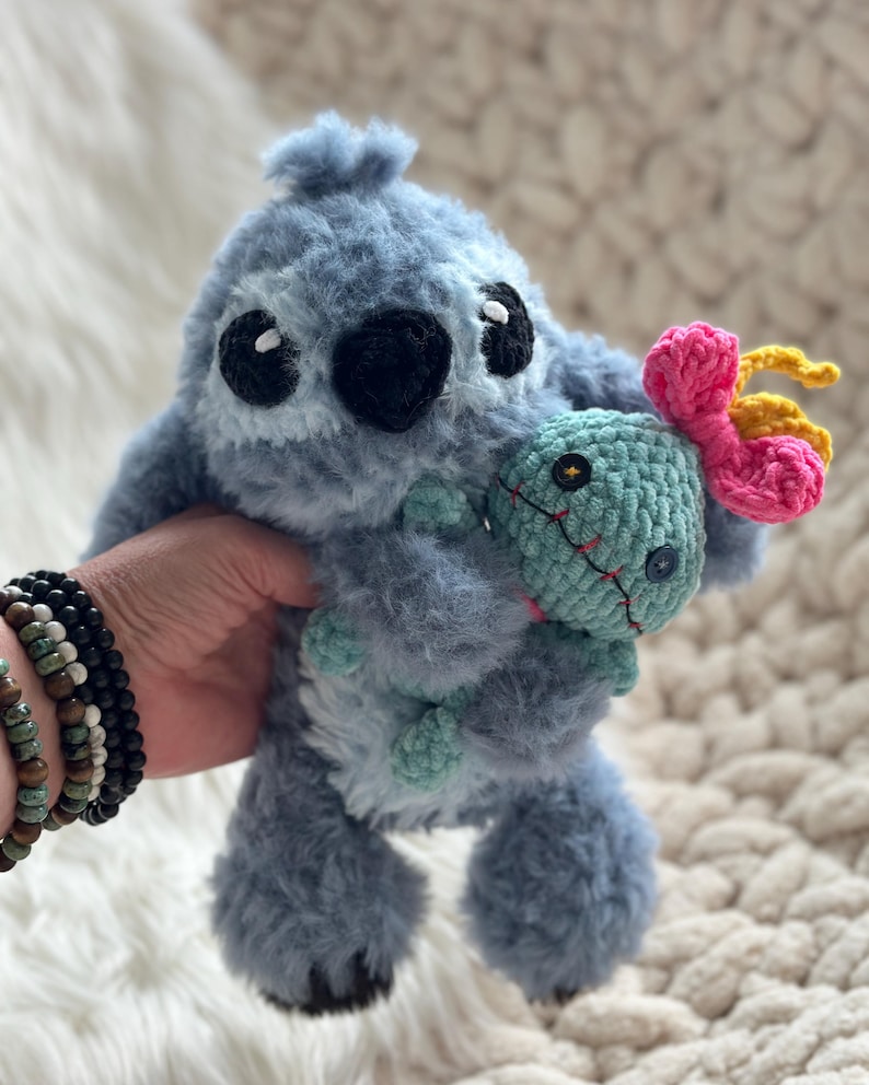 May include: A fuzzy blue crochet plush toy resembling a koala-like creature with large black eyes and a black nose, holding a smaller light green crochet doll that features two dark button eyes, a stitched red mouth, and a bright pink and yellow flower.