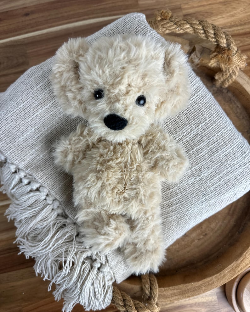 May include: A plush, beige teddy bear with a black nose and eyes, resting on a fringed, light-colored blanket. The bear is positioned in a wooden bowl, with a rope detail visible.