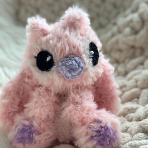 May include: A plush stuffed animal made of soft, fuzzy light pink yarn. It features large, round black eyes with small white highlights, a textured light purple nose, and light purple accents on the bottom of its two front paws.