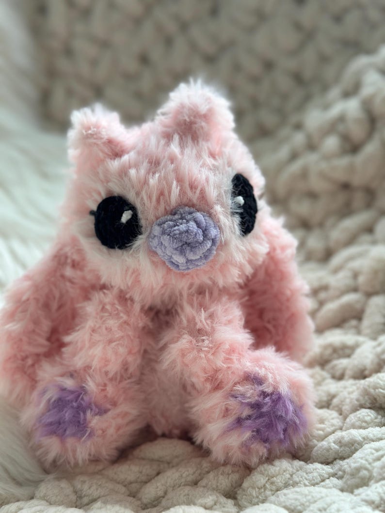 May include: A plush stuffed animal made of soft, fuzzy light pink yarn. It features large, round black eyes with small white highlights, a textured light purple nose, and light purple accents on the bottom of its two front paws.
