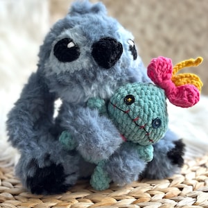 May include: A fuzzy blue plush creature with large ears and black eyes, holding a smaller mint green plush doll. The doll features a button eye, a stitched red mouth, and a bright pink and yellow flower-shaped accessory attached to its head.