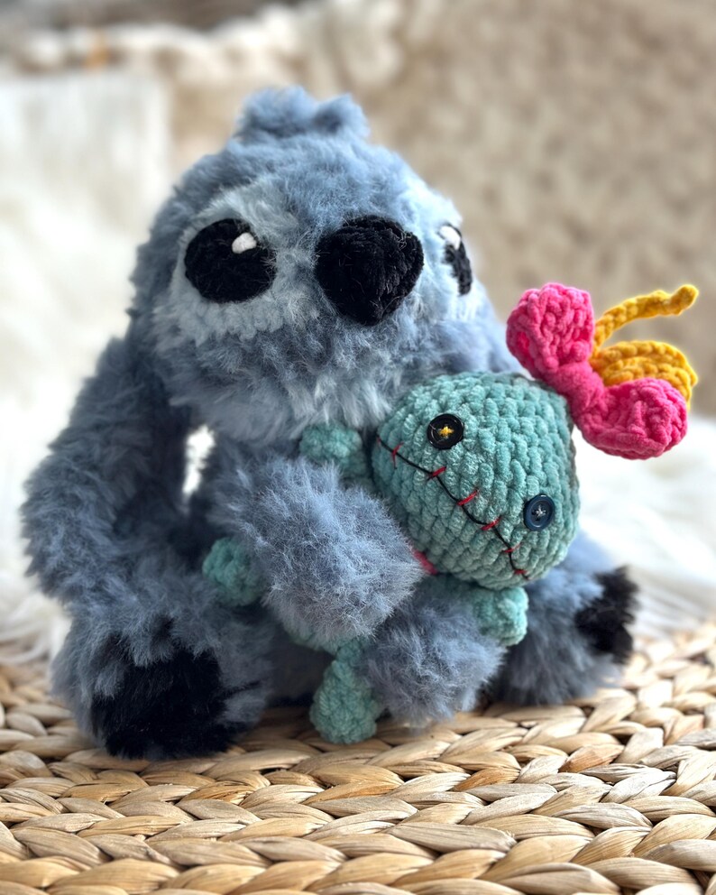 May include: A fuzzy blue plush creature with large ears and black eyes, holding a smaller mint green plush doll. The doll features a button eye, a stitched red mouth, and a bright pink and yellow flower-shaped accessory attached to its head.