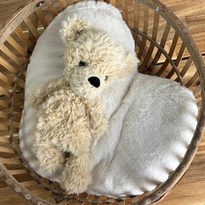 May include: A plush teddy bear with tan fur and black eyes rests on a white, fluffy pillow inside a round, wooden basket. The basket has a woven design and is set against a wooden surface.